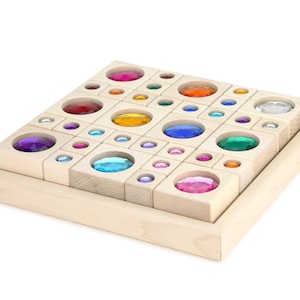 May include: A wooden puzzle with a light wood finish. The puzzle features a grid of various sized openings, each filled with a colorful, gem-like stone. The gems are in shades of red, orange, yellow, green, blue, and purple.