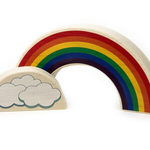 May include: A wooden toy set consisting of a large arched rainbow piece with red, orange, yellow, green, blue, and purple stripes, and a smaller semi-circular wooden piece featuring two light blue outlined clouds on a natural wood grain surface.