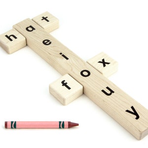 May include: A light-colored wooden spelling toy featuring a long central block with the letters h, a, t, e, i, f, o, u, x, y printed in black, accompanied by a pink crayon. The toy consists of several interlocking wooden pieces.