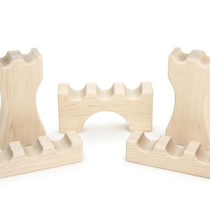 May include: A collection of unfinished wooden wine bottle holders. The holders are light-colored and feature curved slots designed to cradle wine bottles. The set includes two tower-shaped pieces and three rectangular pieces with arched or straight bases.