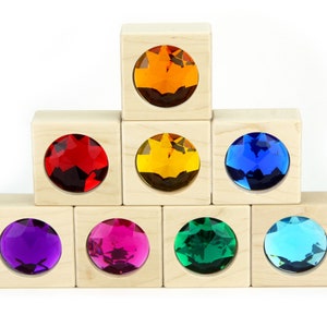 Puede incluir: Un conjunto de ocho bloques de madera con inserciones de vidrio en forma de gemas de colores. Los bloques están dispuestos en forma de pirámide, con el bloque superior que presenta una gema naranja, seguida de gemas rojas, amarillas, azules, moradas, rosas, verdes y turquesas.