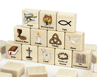 May include: A set of light-colored wooden blocks arranged in a pyramid, each featuring a printed illustration and a corresponding label. The images include an angel, burning bush, fish, bread and wine, candle, trinity, dove, bible, cross, lamb, crown of thorns, and ten commandments.