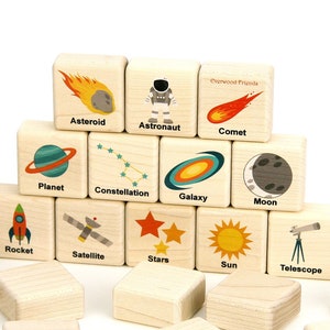 May include: A set of light-colored wooden blocks featuring printed space-themed illustrations and labels including Asteroid, Astronaut, Comet, Planet, Constellation, Galaxy, Moon, Rocket, Satellite, Stars, Sun, and Telescope, with Everwood Friends text.