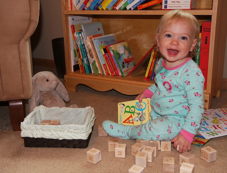 May include: A toddler wearing light blue floral pajamas with pink trim, holding a Dr. Seuss ABC book. Scattered around are wooden alphabet blocks, a dark brown woven basket with a light green polka dot liner, and a plush gray rabbit toy.