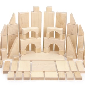 May include: A large collection of natural light-colored wooden building blocks in various geometric shapes, including rectangular prisms, cubes, triangles, and arches, arranged in a symmetrical display on a plain white surface.