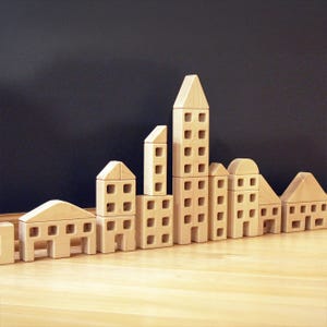 May include: A collection of light-colored wooden building blocks arranged to resemble a city skyline. The set includes various shapes such as rectangular towers, houses with triangular roofs, and structures with arched tops, all featuring square cutouts.