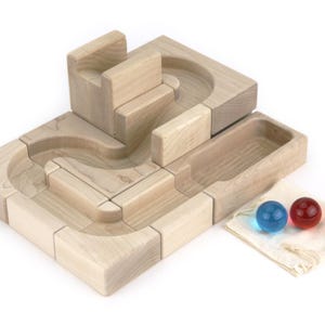 May include: A light-colored wooden marble run set featuring interlocking blocks with carved tracks, a rectangular storage tray, and two translucent glass marbles in blue and red, accompanied by a small off-white fabric drawstring pouch.