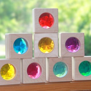 May include: A set of seven light-colored wooden blocks arranged in a pyramid. Each block features a circular cutout containing a faceted, translucent acrylic gem in a different color, including red, blue, yellow, purple, pink, teal, and green.