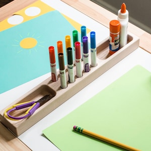 May include: A wooden organizer with compartments for markers, glue sticks, and scissors. The organizer is filled with colorful markers, a glue stick, and a pair of purple scissors. There are also sheets of colored paper and a yellow pencil on the table.