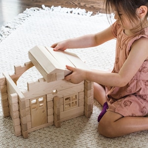 May include: A light-colored wooden toy cabin construction set featuring interlocking logs, a rectangular door with a window, a square window frame, and a removable roof section. The structure is built from smooth, natural wood blocks.
