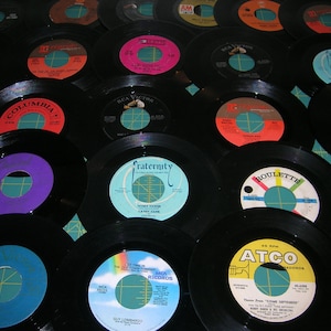May include: A collection of vintage vinyl records in various colors, including black, white, red, blue, and yellow. The records are arranged in a grid pattern on a green surface. Some of the records have labels with text, including "Columbia", "RCA Victor", "Fraternity", "Roulette", and "Atco".