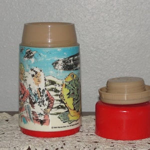 Puede incluir: Un termo vintage marr&oacute;n y rojo con un gr&aacute;fico de personajes de la pel&iacute;cula 'La Criatura de la Laguna Negra'. El termo tiene una tapa marr&oacute;n y una base roja. El gr&aacute;fico presenta a una mujer con un vestido blanco, un hombre con una camisa azul y una criatura de la Laguna Negra.