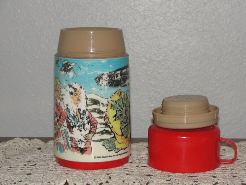 Puede incluir: Un termo vintage marr&oacute;n y rojo con un gr&aacute;fico de personajes de la pel&iacute;cula 'La Criatura de la Laguna Negra'. El termo tiene una tapa marr&oacute;n y una base roja. El gr&aacute;fico presenta a una mujer con un vestido blanco, un hombre con una camisa azul y una criatura de la Laguna Negra.