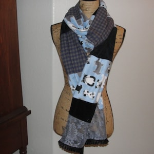 May include: A patchwork scarf displayed on a mannequin. The scarf features various patterned fabrics, including blue plaid, black, and a light blue fabric with animal prints. The bottom edge has a black lace trim.