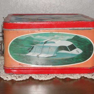 Puede incluir: Fiambrera de metal roja con un dise&ntilde;o de nave espacial blanca y el texto "S.F.".