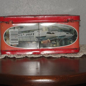 Puede incluir: Fiambrera de metal roja con una imagen de un tren futurista blanco en la parte delantera. El tren est&aacute; en un t&uacute;nel con un c&oacute;digo de barras en el lateral.