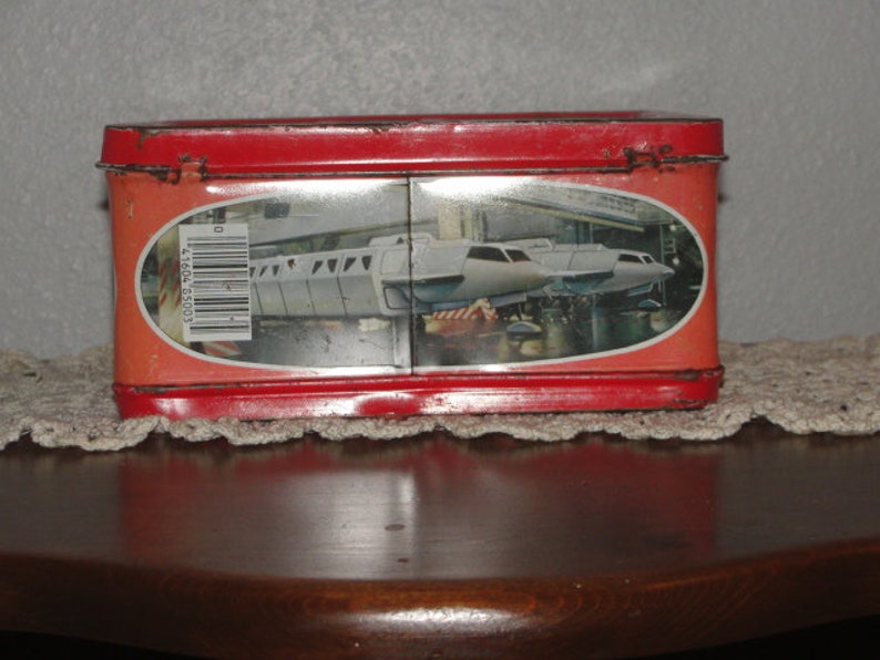 Puede incluir: Fiambrera de metal roja con una imagen de un tren futurista blanco en la parte delantera. El tren est&aacute; en un t&uacute;nel con un c&oacute;digo de barras en el lateral.