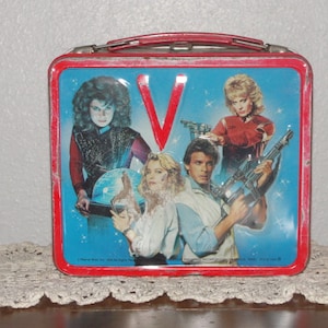 Puede incluir: Lonchera vintage de metal con el elenco del programa de televisi&oacute;n de los a&ntilde;os 80 "V". La lonchera es roja con un interior azul y presenta el logotipo del programa, "V", en rojo. Los personajes se representan en poses de acci&oacute;n, sosteniendo armas y con una mirada decidida.