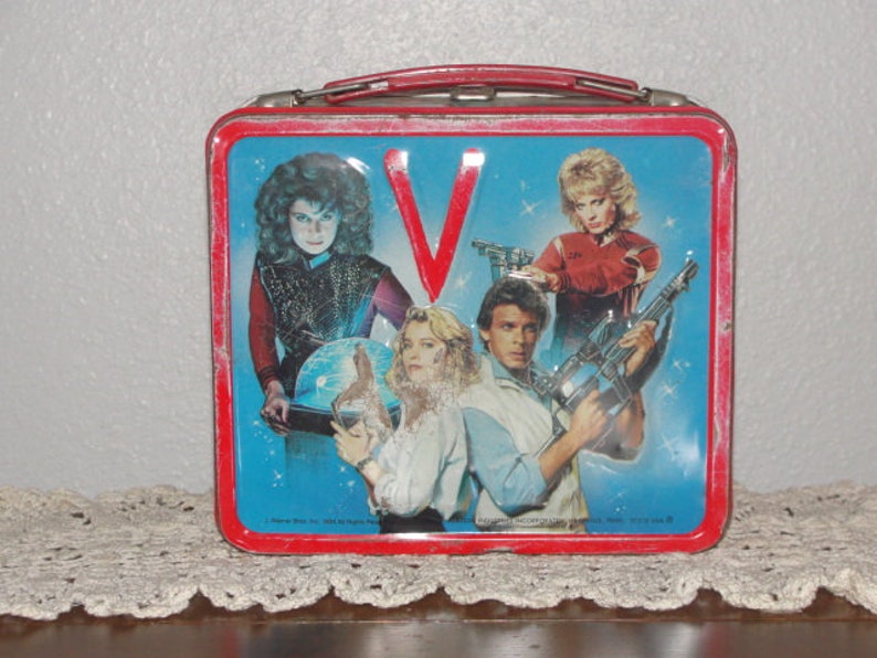Puede incluir: Lonchera vintage de metal con el elenco del programa de televisi&oacute;n de los a&ntilde;os 80 "V". La lonchera es roja con un interior azul y presenta el logotipo del programa, "V", en rojo. Los personajes se representan en poses de acci&oacute;n, sosteniendo armas y con una mirada decidida.