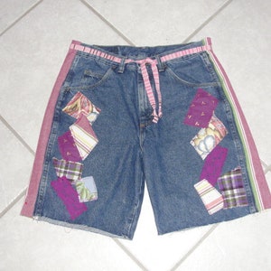 May include: A pair of blue denim shorts with pink and green trim. The shorts have multiple patches of fabric sewn on in a patchwork design. The patches are in shades of purple, pink, and white.