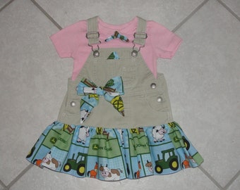 Puede incluir: Un vestido de peto beige con una camisa rosa de manga corta debajo. El vestido de peto tiene un lazo azul y verde y una falda con volantes con un estampado de animales de granja y tractores John Deere.