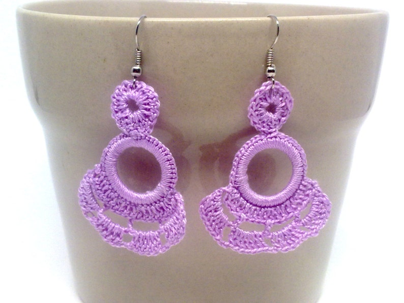 May include: A pair of lavender crocheted earrings with a circle design. The earrings are hanging from silver hooks.