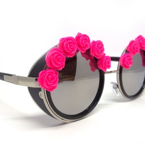 May include: Round black sunglasses with a silver metal frame and pink plastic rose embellishments around the lenses.