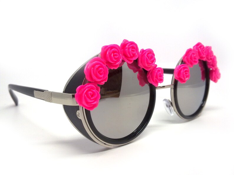 May include: Round black sunglasses with a silver metal frame and pink plastic rose embellishments around the lenses.