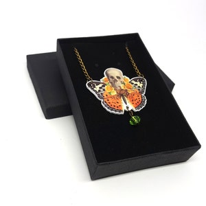 May include: A necklace with a butterfly pendant featuring a skull with flowers. The butterfly is orange, black, and white. The pendant is hanging from a bronze chain and has a green crystal bead at the bottom.