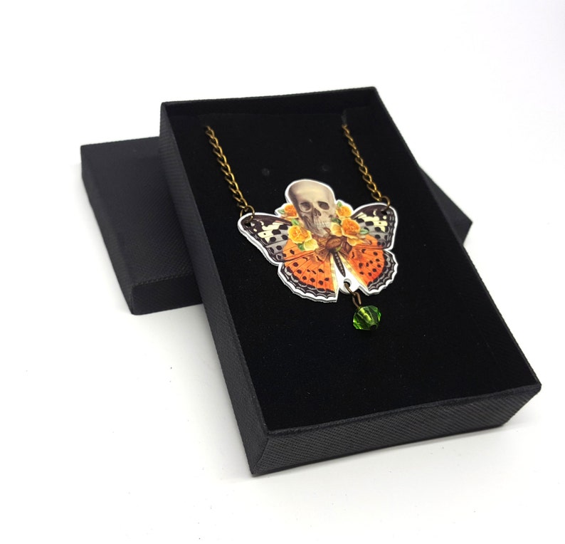 May include: A necklace with a butterfly pendant featuring a skull with flowers. The butterfly is orange, black, and white. The pendant is hanging from a bronze chain and has a green crystal bead at the bottom.