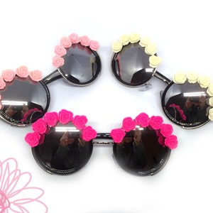 May include: Four pairs of round sunglasses with decorative flower accents. Two pairs have pink flowers, one pair has white flowers, and one pair has hot pink flowers.