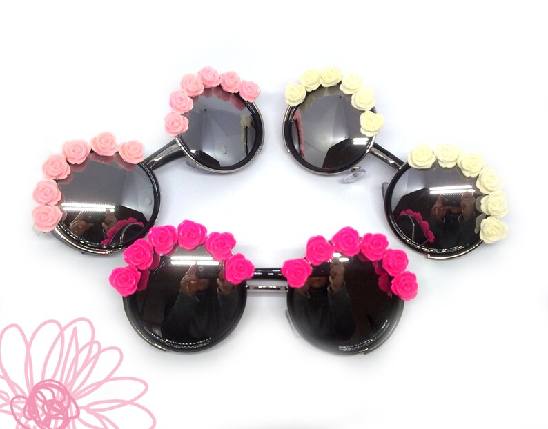 May include: Four pairs of round sunglasses with decorative flower accents. Two pairs have pink flowers, one pair has white flowers, and one pair has hot pink flowers.