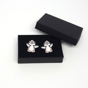 May include: A pair of cufflinks in a black box. Each cufflink features a cartoon penguin with black and white wings and a white body. The penguin has black eyes and an orange beak.