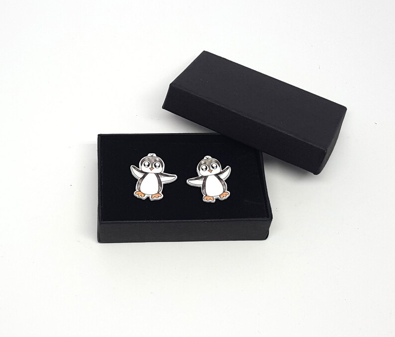 May include: A pair of cufflinks in a black box. Each cufflink features a cartoon penguin with black and white wings and a white body. The penguin has black eyes and an orange beak.