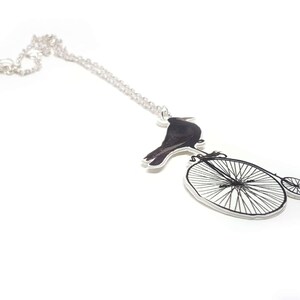 May include: A silver chain necklace with a pendant featuring a black crow perched on a vintage bicycle with a large front wheel and a smaller back wheel.