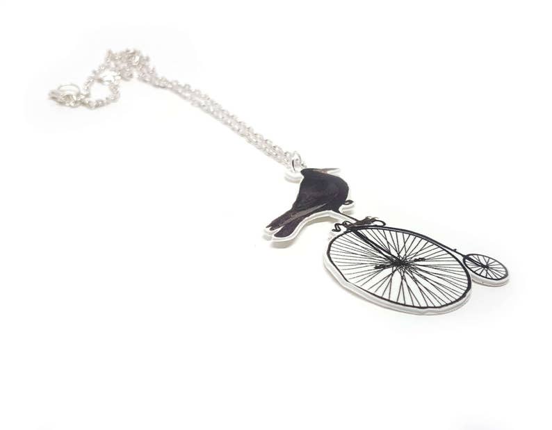 May include: A silver chain necklace with a pendant featuring a black crow perched on a vintage bicycle with a large front wheel and a smaller back wheel.