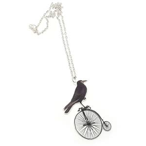 May include: A silver chain necklace with a pendant featuring a black crow riding a vintage bicycle with a single large wheel.