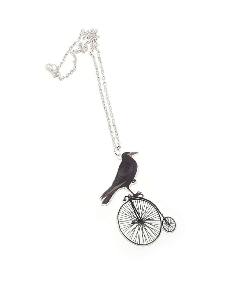 May include: A silver chain necklace with a pendant featuring a black crow riding a vintage bicycle with a single large wheel.
