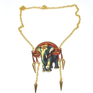Puede incluir: Collar de cadena dorada con un colgante que presenta una ilustración colorida de un elefante en un palanquin rojo y dorado. El colgante tiene tres púas doradas que cuelgan de él.