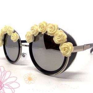 May include: Round black sunglasses with a silver metal frame and a row of white plastic flowers across the top.