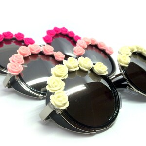 May include: Three pairs of round sunglasses with black frames and black lenses. Each pair has a row of plastic flowers around the lenses. The flowers are pink, light pink, and white.