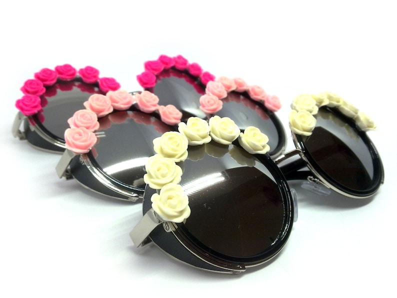 May include: Three pairs of round sunglasses with black frames and black lenses. Each pair has a row of plastic flowers around the lenses. The flowers are pink, light pink, and white.