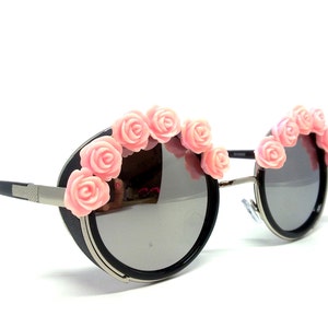 May include: Round black sunglasses with a silver frame and pink flower accents.