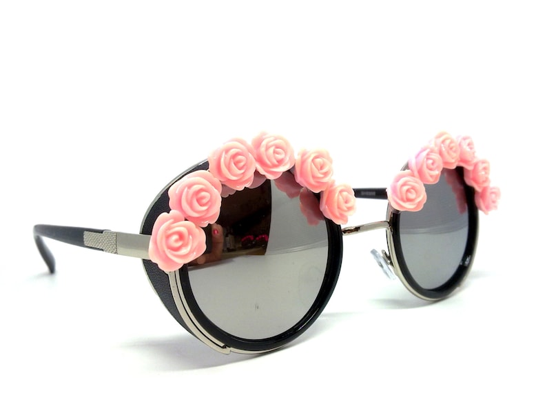 May include: Round black sunglasses with a silver frame and pink flower accents.