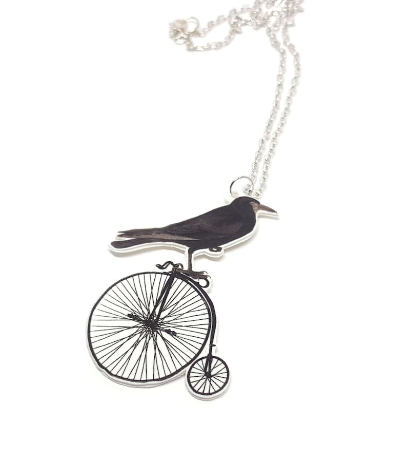 May include: A silver chain necklace with a black crow perched on a vintage bicycle pendant. The crow is facing right and the bicycle has a large front wheel and a smaller back wheel.