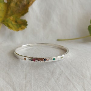 May include: A silver bangle bracelet with a hammered finish and five small gemstones set in the metal. The gemstones are red, pink, blue, green, and clear.
