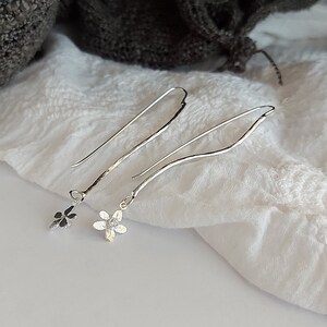 May include: A pair of silver dangle earrings with a delicate flower charm. The earrings are long and thin, with a slightly curved shape. The flower charms are small and have a simple design.