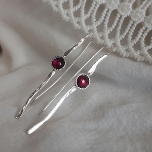 Könnte beinhalten: Ein Paar silberne Ohrringe mit einem tief roten Edelstein in jedem. Die Ohrringe haben eine gehämmerte Textur und ein langes, dünnes Design.