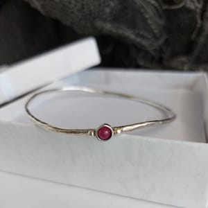 Peut inclure: Un bracelet jonc en argent avec une texture martelée et une petite pierre précieuse rouge ronde au centre. Le bracelet est présenté dans une boîte blanche, suggérant un bijou.