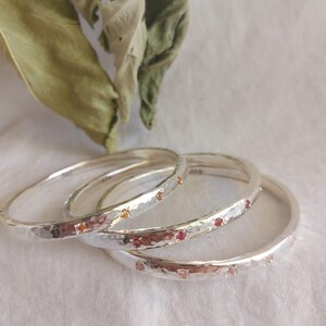 May include: Three silver bangles with hammered texture and small red and orange gemstones set into the metal.